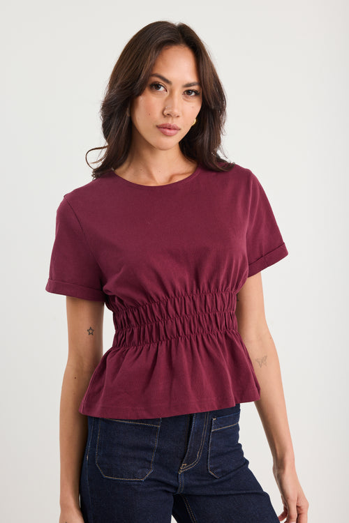 Woman wearing a maroon peplum top and dark jeans against a white background