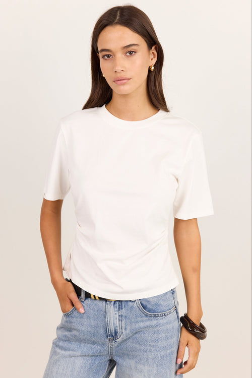 A woman modeling a plain white short-sleeve crewneck t-shirt paired with light-wash blue jeans, standing against a solid off-white studio background.