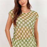 A woman modeling a sheer olive green mesh dress with a crisscross grid pattern over a light beige underlayer, set against a plain off-white studio background.