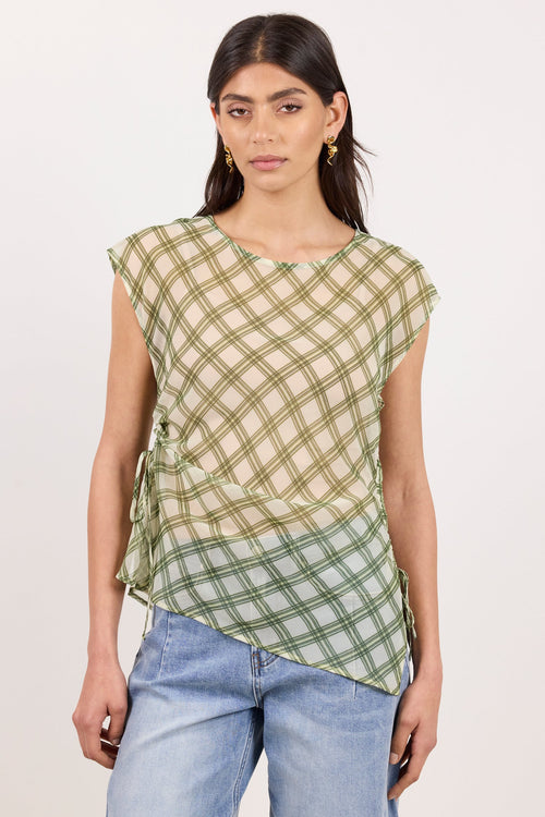A woman modeling a sleeveless, asymmetrical top with a green and beige plaid pattern, paired with light-wash blue jeans, against a plain off-white studio background.