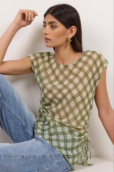 A woman sits on a white floor against a white wall wearing a green and white plaid sleeveless top with side ruffles and light blue jeans.
