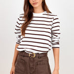A woman modeling a white long-sleeve knit top with dark brown horizontal stripes, tucked into dark brown denim shorts with a matching belt, against a plain light gray studio background.