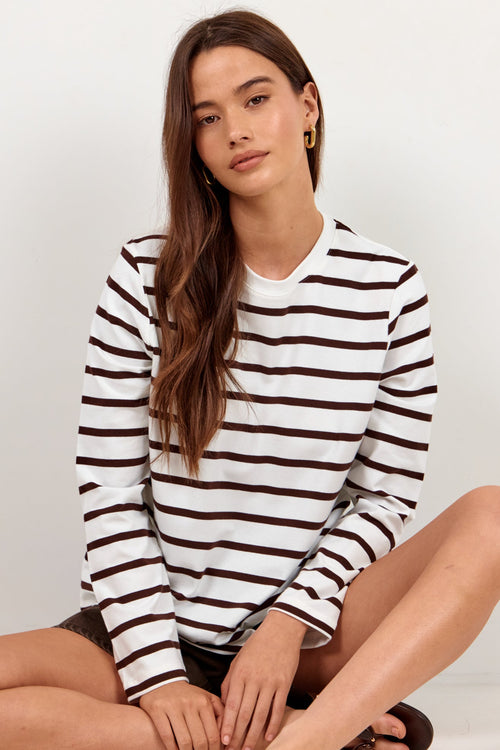 A woman sits cross-legged on a white studio floor wearing a white long-sleeve top with dark brown horizontal stripes, dark shorts, and black sandals.