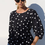 A woman wearing a black long-sleeve blouse with white polka dots and blue jeans, standing against a white wall in bright sunlight.
