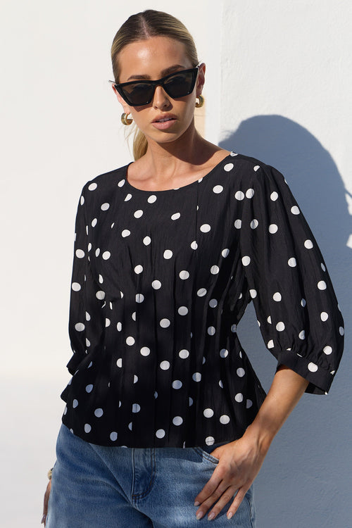 A woman wearing a black long-sleeve blouse with white polka dots and blue jeans, standing against a white wall in bright sunlight.
