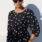 A woman wearing a black long-sleeve blouse with white polka dots and blue jeans, standing against a white wall in bright sunlight.