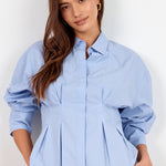 A woman wearing a light blue button-up blouse with dramatic puffed sleeves and a gathered peplum waist, paired with black pants, against a plain white background.