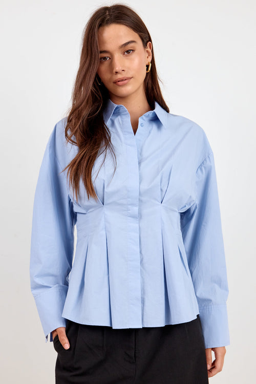 A woman modeling a light blue long-sleeve button-up blouse with a peplum waist and front pleats, paired with black trousers, against a plain light gray studio background.