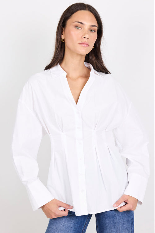 A woman wearing a white long-sleeve button-up blouse with a gathered waist and peplum-like drape, paired with blue jeans, against a plain light gray studio background.