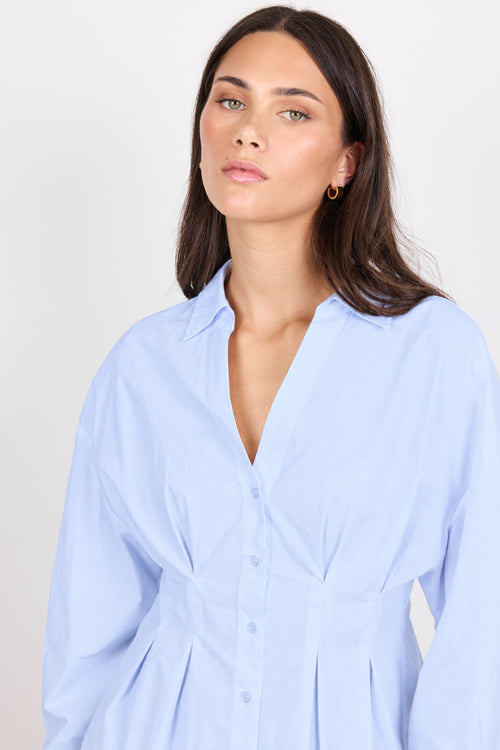 A woman wearing a light blue button-up shirt dress with a V-neck and gathered waist details, photographed against a plain light gray studio background.