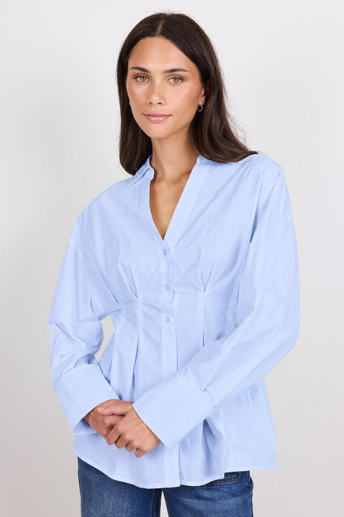 A woman modeling a light blue long-sleeve button-up blouse with a gathered peplum-style waist, paired with blue jeans, against a plain light gray studio background.