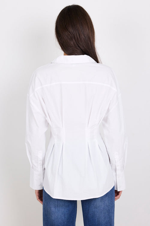 A woman viewed from the back wearing a white long-sleeve button-up shirt with gathered detailing at the waist and blue jeans, set against a plain light gray background.