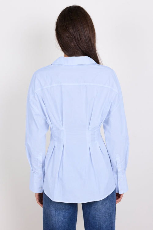 A woman viewed from the back wearing a light blue long-sleeve button-up shirt with a peplum waist and dark blue jeans, set against a plain off-white background.
col_pattern