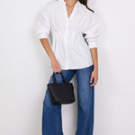A woman modeling a white oversized button-up shirt with blue wide-leg jeans, black sandals, and a black handbag against a plain white studio background.
