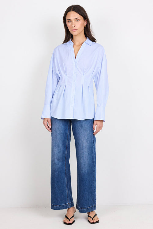 A woman modeling a light blue long-sleeve button-up shirt with a gathered waist, paired with wide-leg blue jeans and black flip-flops, standing against a plain white studio background.