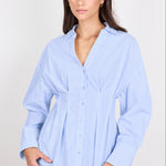A woman modeling a light blue long-sleeve button-up shirt with a gathered peplum waist, paired with blue jeans, against a plain light gray background.