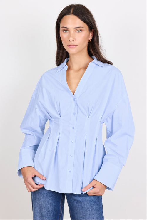 A woman modeling a light blue long-sleeve button-up shirt with a gathered peplum waist, paired with blue jeans, against a plain light gray background.