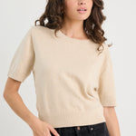 Woman wearing a beige sweater and black pants against a white background
