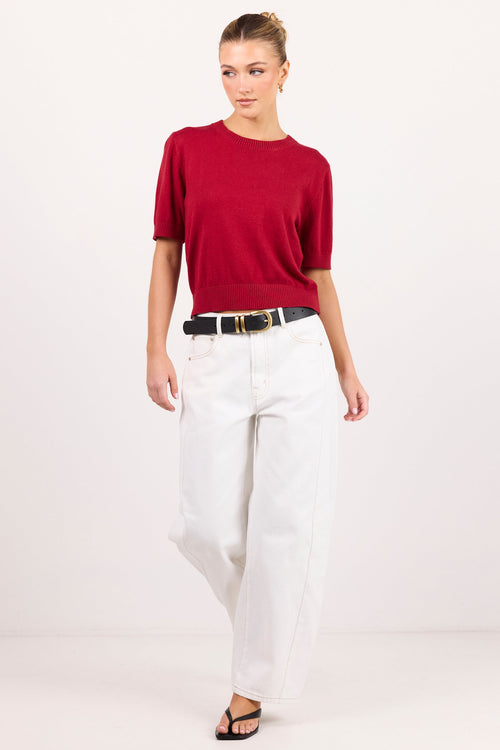 A woman modeling a short-sleeve crewneck knit top in deep red, paired with white wide-leg pants, a black belt, and black sandals, against a plain white studio background.
