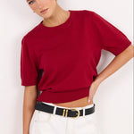A woman modeling a short-sleeve knit top in deep red, paired with white pants and a black belt with gold hardware, against a plain white studio background.