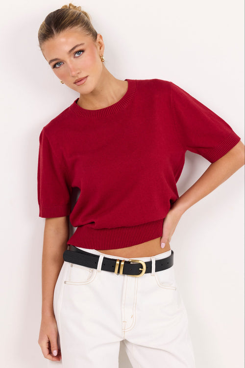 A woman modeling a short-sleeve knit top in deep red, paired with white pants and a black belt with gold hardware, against a plain white studio background.
