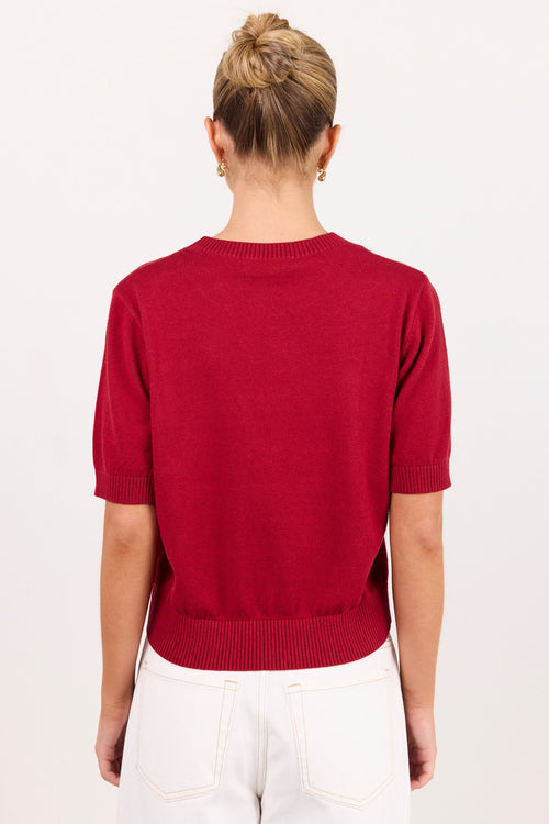 A woman viewed from behind wearing a short-sleeve knit sweater in deep red and white pants, standing against a plain white studio background.