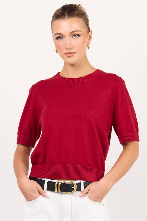 A woman modeling a short-sleeve crewneck knit top in deep red, paired with white wide-leg jeans, a black belt, and black sandals, standing against a plain white studio background.