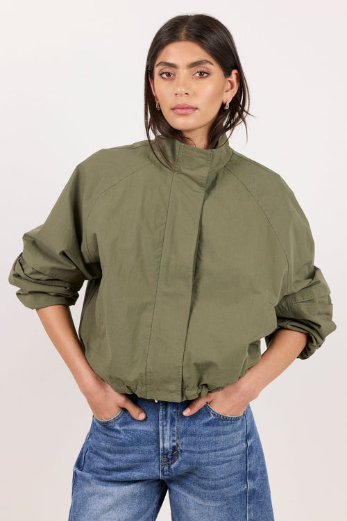 A woman modeling an olive green lightweight jacket with a high stand-up collar and voluminous sleeves, paired with blue jeans, against a plain light background.