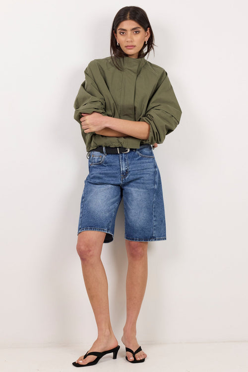 A model stands against a plain white studio background wearing an olive green cropped jacket, blue denim shorts, and black heeled sandals.