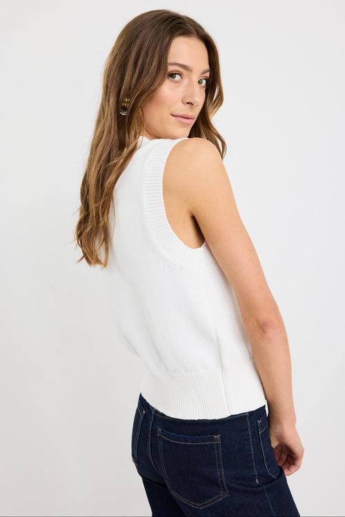 Woman wearing an ivory sleeveless knit vest top with gold button front closure and a crew neckline, styled with dark indigo jeans.
