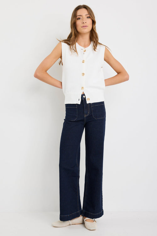 Woman wearing an ivory sleeveless knit vest top with gold button front closure and a crew neckline, styled with dark indigo jeans.