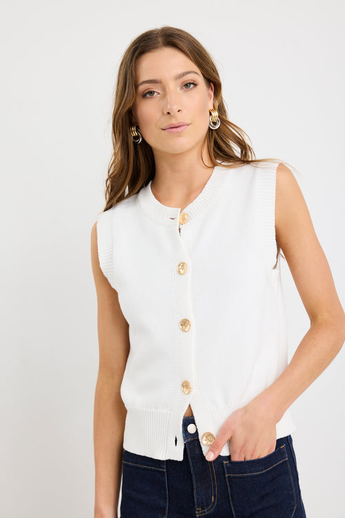 Woman wearing an ivory sleeveless knit vest top with gold button front closure and a crew neckline, styled with dark indigo jeans.