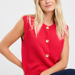 Woman wearing a sleeveless red knit vest with gold buttons down the front, styled with dark denim and loose blonde waves.
