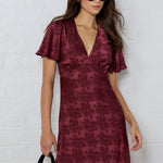 Woman wearing a burgundy patterned dress against a white brick wall.