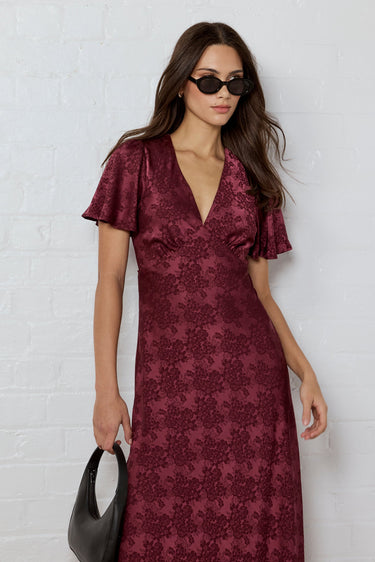 Woman wearing a burgundy patterned dress against a white brick wall.