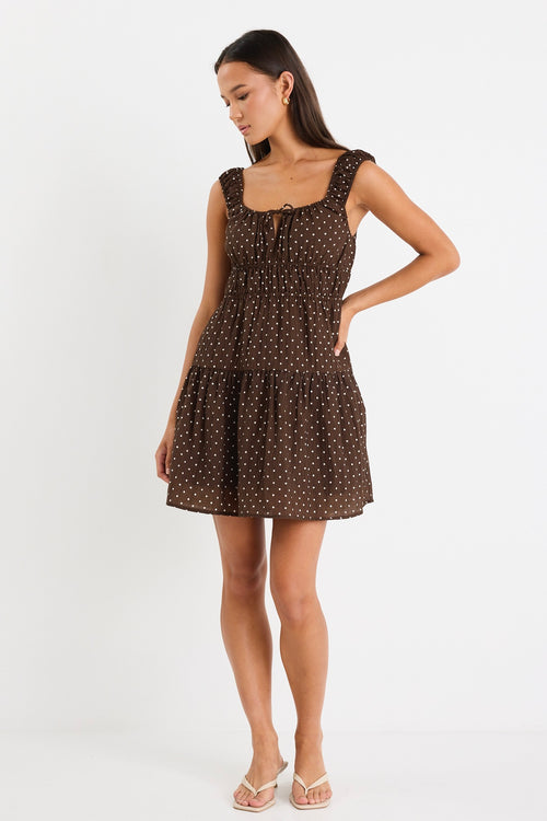Model wears a sleeveless brown mini dress with white polka dots, featuring a tiered skirt, ruffled straps, and a tie-front detail. 