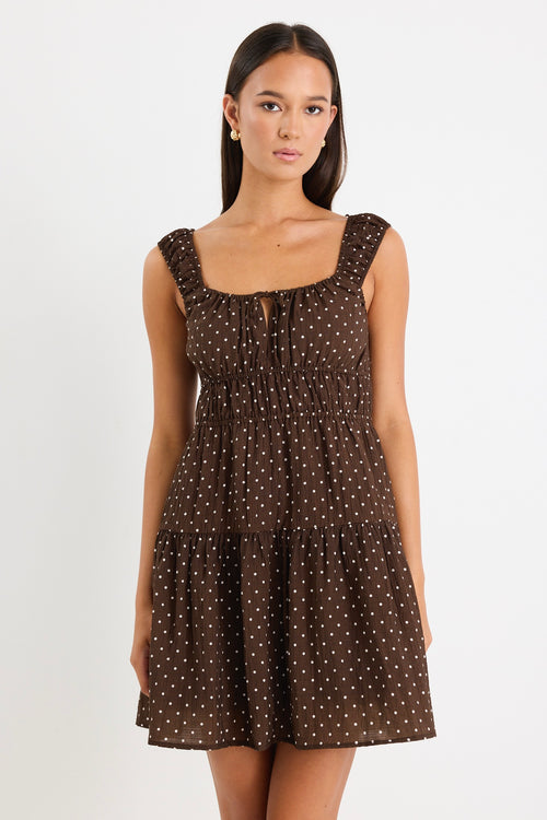 Model wears a sleeveless brown mini dress with white polka dots, featuring a tiered skirt, ruffled straps, and a tie-front detail. 