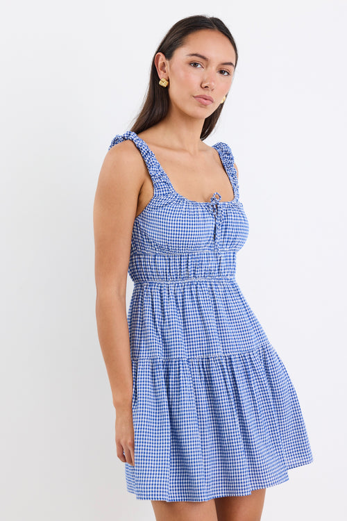 A sleeveless electric blue and white gingham mini dress with tiered detailing, tie-front feature, and a flattering fit. Fully lined for comfort, made from a 50% Cotton and 50% Viscose blend.