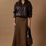 Woman wearing a brown leather jacket and patterned skirt holding a matching handbag against a brown background