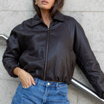 Woman wearing a black leather jacket and blue jeans against a gray wall.