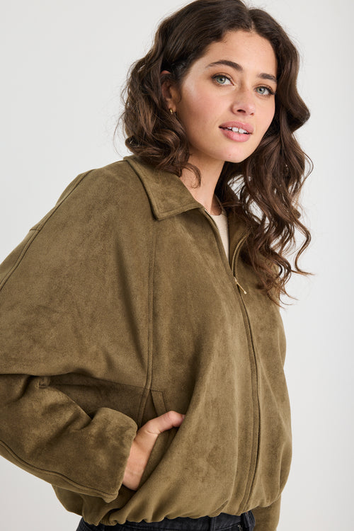 Woman wearing a khaki mock suede zip-up jacket with a soft, textured finish and a relaxed fit, styled over a beige knit top and black jeans.