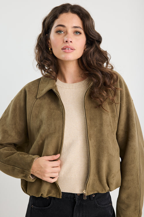 Woman wearing a khaki mock suede zip-up jacket with a soft, textured finish and a relaxed fit, styled over a beige knit top and black jeans.