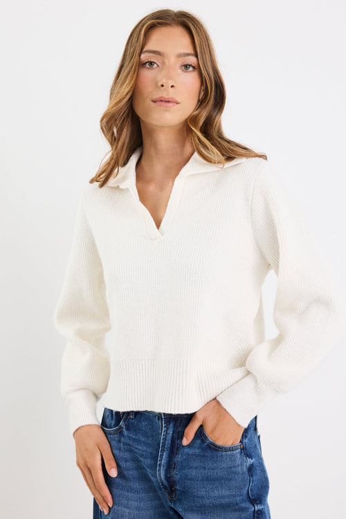 Woman wearing a white sweater and blue jeans against a plain background