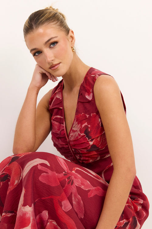 A woman wearing a sleeveless deep V-neck dress with a red and pink floral pattern, posing against a plain white background.