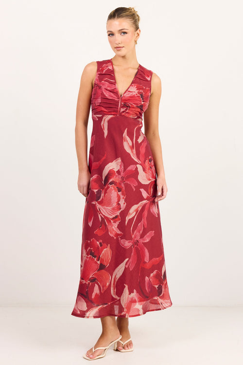 A woman modeling a sleeveless red maxi dress with a large pink and white floral print, standing against a plain white studio background.