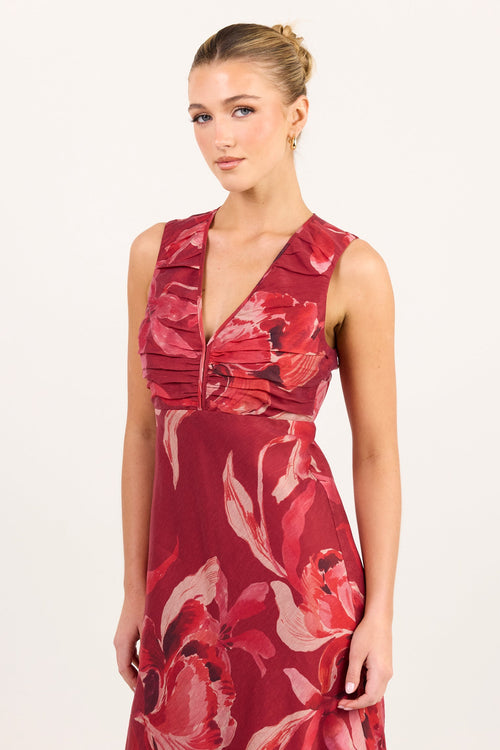 A woman modeling a sleeveless red maxi dress with a large floral print, a deep V-neckline, and a gathered waist detail, set against a plain off-white studio background.