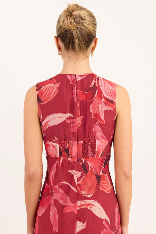 A woman viewed from the back wearing a sleeveless red dress with a pink and coral floral print and a gathered waist detail, against a plain white background.