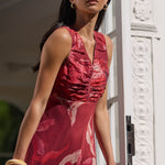 A woman wearing a sleeveless red floral maxi dress and black sunglasses stands on a balcony with a black metal railing, leaning on the railing against a white building facade.