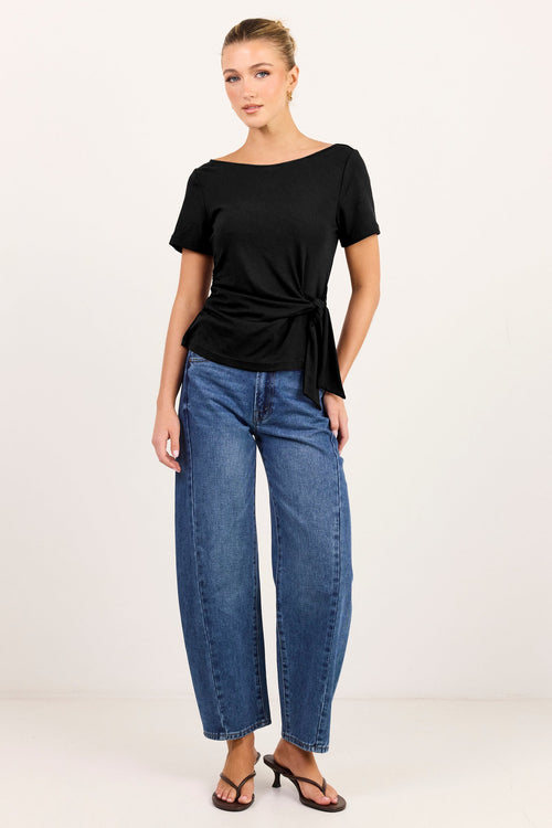 A woman modeling a black short-sleeve top with a side tie detail and blue wide-leg jeans against a plain white studio background.