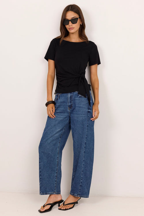 A woman modeling a black short-sleeve t-shirt with a twisted knot detail at the hem, paired with wide-leg blue jeans and black sandals, standing against a plain off-white studio background.